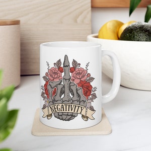 May include: White ceramic mug featuring a gray skeletal hand giving the middle finger, surrounded by pink and red roses. A banner below the hand reads "NEGATIVITY". The mug is on a beige coaster.