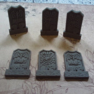 May include: Six small, grey, tombstone-shaped decorations with different designs. The designs include a bat, a spider web, and the words "RIP".
