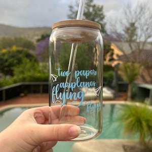 May include: A clear glass reusable drinking cup with a bamboo lid and a straw. The cup has a blue and white design that says "two paper airplanes flying".
