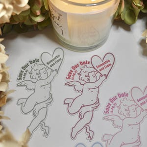 Pode incluir: Uma vela acesa num frasco de vidro junto a vários autocolantes. Os autocolantes apresentam um desenho em forma de coração com um anjo e o texto "Save Our Date" e os nomes "Samantha and Liam" com a data 12.09.27.
