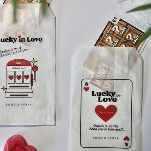 以下が含まれることがあります： 「Lucky in Love」の文字とイラストが描かれた白い紙袋が2つ。1つはスロットマシンのデザイン、もう1つはトランプのデザイン。それぞれスクラッチくじが入っています。赤いハート型のロリポップが表面に置かれています。