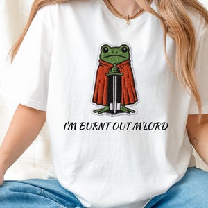 May include: White t-shirt featuring a green frog wearing a red cape and holding a sword. The text "I'M BURNT OUT M'LORD" is printed below the graphic. The shirt is worn by a person wearing blue jeans.