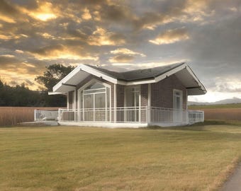 May include: A single-story house with dark brown brick walls, a white gabled roof, and white trim. The structure features large glass windows, a white metal railing surrounding a porch, and a white door, set against a cloudy sunset sky.
