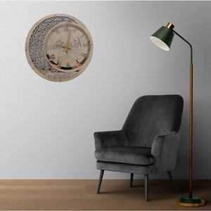 May include: A wooden clock with a crescent moon design and Arabic numerals. The clock face is white with black Roman numerals and a gold-colored frame. The clock is hanging on a white wall with a gray armchair and a green floor lamp.