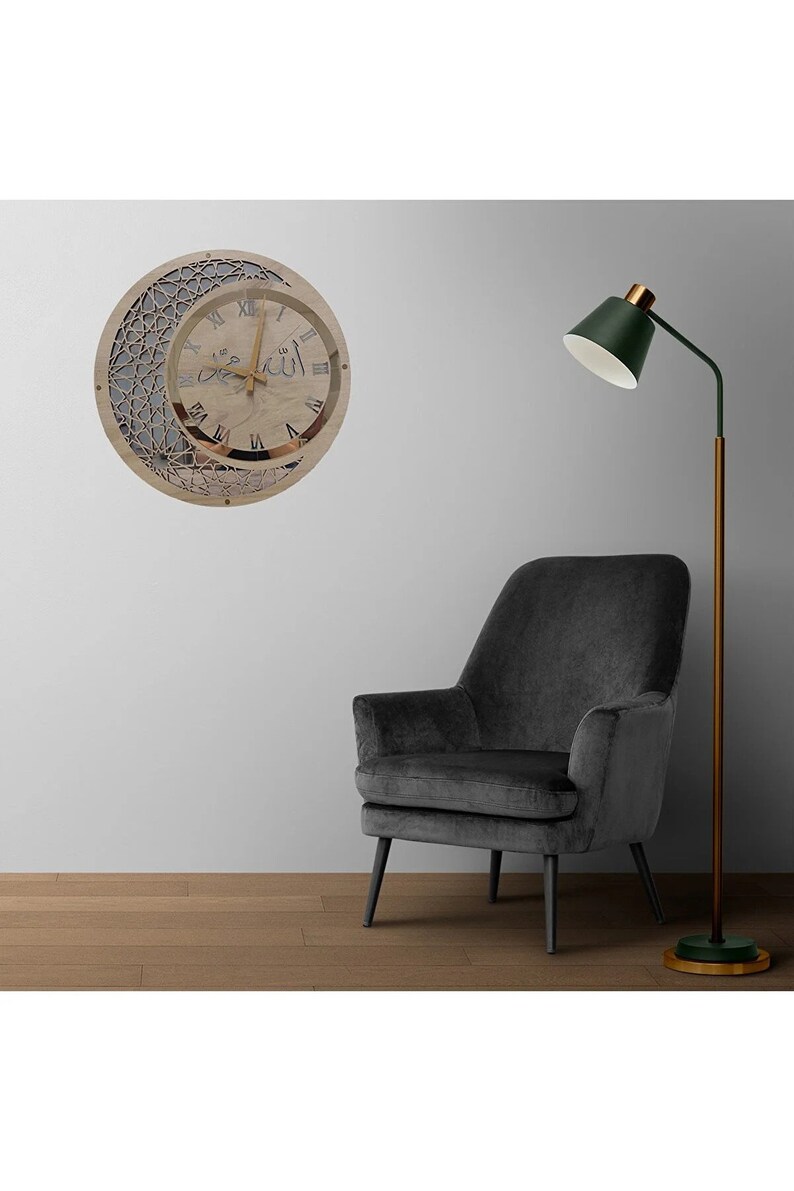 May include: A wooden clock with a crescent moon design and Arabic numerals. The clock face is white with black Roman numerals and a gold-colored frame. The clock is hanging on a white wall with a gray armchair and a green floor lamp.