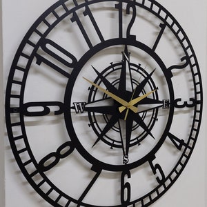 Könnte beinhalten: Schwarze Wanduhr aus Metall mit römischen Ziffern und einem Kompass-Design in der Mitte. Die Zeiger der Uhr sind golden.