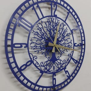 May include: A blue metal wall clock with a tree of life design. The clock has Roman numerals and gold hands. It's mounted on a white wall. The clock's diameter is approximately 30 cm.