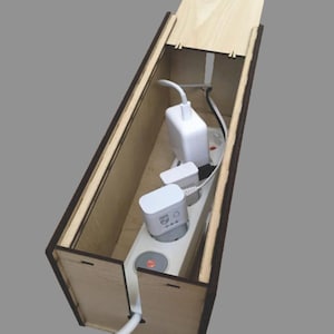Puede incluir: Una caja de madera con tapa, diseñada para contener y ocultar una regleta y cables. La caja tiene una ranura para que los cables pasen. La regleta es blanca y tiene tres enchufes y un interruptor.