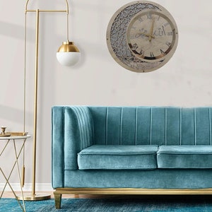 May include: A wooden wall clock with a crescent moon and Arabic calligraphy design. The clock face has Roman numerals and a gold-colored frame. The clock is hanging on a white wall above a teal blue sofa.