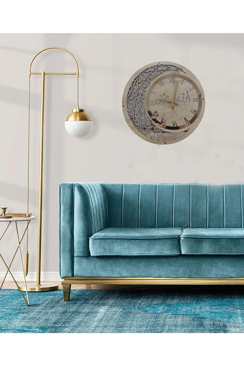 May include: A wooden wall clock with a crescent moon and Arabic calligraphy design. The clock face has Roman numerals and a gold-colored frame. The clock is hanging on a white wall above a teal blue sofa.