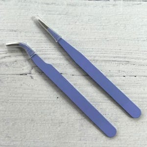 May include: Two pairs of purple tweezers with clear tips. The tweezers are on a white wooden surface.