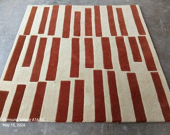Puede incluir: Una alfombra rectangular con un patrón de alto contraste de rayas verticales gruesas de color naranja óxido sobre un fondo de color crema sólido. Las rayas están dispuestas en un diseño escalonado y no uniforme en toda la superficie.