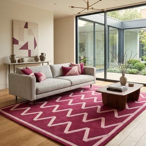 Puede incluir: Un salón moderno con un sofá gris y cojines rosas, una alfombra magenta con un diseño en zigzag blanco y una mesa de centro de madera. Una lámpara de araña dorada cuelga encima. El logotipo de TheFineRugs está en la esquina inferior derecha.