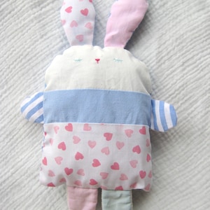 Op de afbeelding: Een handgemaakt knuffelkonijn met een witte body, blauwe en roze accenten. De oren van het konijn zijn roze en bedrukt met roze hartjes. De body van het konijn heeft een blauwe streep en een gedeelte met roze hartjes. Het speelgoed heeft een gestikt gezicht.