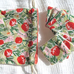 Op de afbeelding: Een trekkoordtas met een bloemenprint van rode en roze bloemen op een witte achtergrond. De tas is gemaakt van stof en heeft een roze bies. De tas is open en er zitten verschillende kleine stoffen vierkanten in de tas. De vierkanten zijn ook bedrukt met rode en roze bloemen op een witte achtergrond.