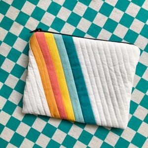Può includere: Pochette trapuntata bianca con un design a righe arcobaleno. Le strisce arcobaleno includono tonalità di corallo, arancione, giallo, azzurro e turchese. La pochette ha una cerniera nera ed è appoggiata su uno sfondo a scacchi turchese e bianco.