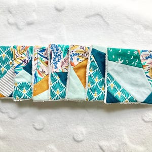 Op de afbeelding: Een rij kleurrijke, bedrukte stoffen vierkanten. De stoffen hebben bloemen-, geometrische en abstracte patronen in tinten teal, goud en roze. Deze vierkanten maken waarschijnlijk deel uit van een groter textielproject.