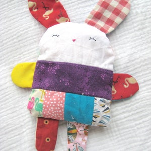 Puede incluir: Un juguete de conejo de patchwork con una cara blanca, tela morada, azul, amarilla, rosa y roja. El conejo tiene ojos bordados y una nariz rosa.