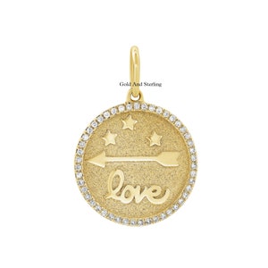 Może przedstawiać: Złoty naszyjnik z zawieszką w kształcie okrągłego wisiorka z przedstawioną złotą strzałą, trzema złotymi gwiazdami i napisem "love" w ozdobnym piśmie. Wisiorek jest otoczony kółkiem z małych, przezroczystych diamentów.