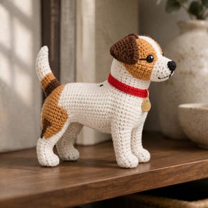 May include: A crocheted Jack Russell Terrier dog figurine. The dog is white with brown and tan markings, a red collar, and a gold tag. The dog is standing on a wooden shelf.