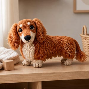 Peut inclure: Un jouet en forme de chien teckel au crochet, dans les tons marron, beige et crème. Le chien à poils longs a un visage détaillé avec des yeux noirs et un museau crème. Le jouet est posé sur une surface en bois.
