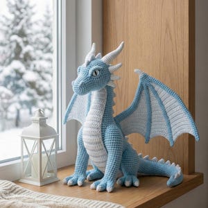 Könnte beinhalten: Hellblau-weiß gehäkelte Drachenfigur mit großen Flügeln, Hörnern und einem langen Schwanz. Der Drache sitzt auf einer Holzfensterbank neben einer weißen Laterne. Der Hintergrund zeigt eine verschneite Winterszene.