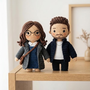 Puede incluir: Dos muñecos de ganchillo hechos a mano, una pareja, están uno al lado del otro. La muñeca femenina lleva gafas, un vestido azul, un abrigo gris y un bolso marrón. El muñeco masculino lleva una chaqueta azul marino, una camisa blanca y pantalones negros. Ambos muñecos tienen pelo castaño y se dan la mano.