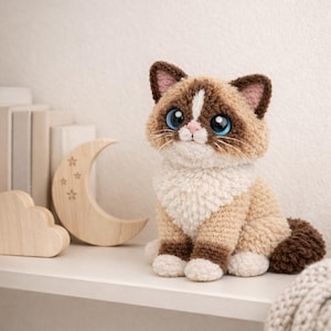 May include: A handmade crochet cat doll with brown and white fur, large blue eyes, and a pink nose. The cat is sitting on a white shelf next to wooden decorative shapes, including a moon and cloud. The cat has a fluffy tail.