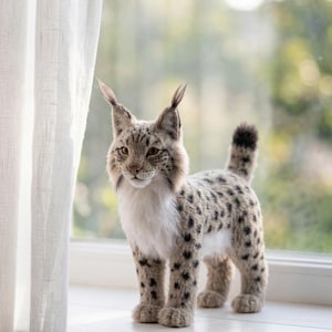 Op de afbeelding: Een pluche lynx figuur met een gevlekte vacht, staand op een witte vensterbank. De lynx heeft gepluimde oren, een witte borst en een korte, zwartgepuntte staart. Een doorschijnend wit gordijn is links zichtbaar.