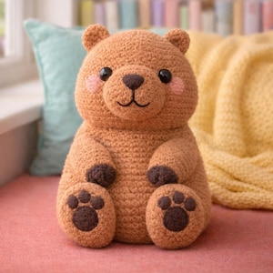 May include: A crocheted teddy bear in shades of brown. The bear has dark brown paws and a snout, with pink cheeks and black eyes. The bear is sitting on a pink surface, with a blue cushion and yellow blanket in the background.
