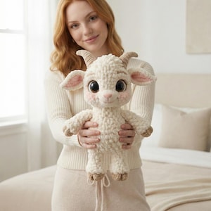 以下が含まれることがあります： 大きな瞳、ピンクの頬、小さな角が特徴の、クリーム色のぬいぐるみの子ヤギ。柔らかく、質感のある外観で、ニュートラルな背景の前に抱えられています。