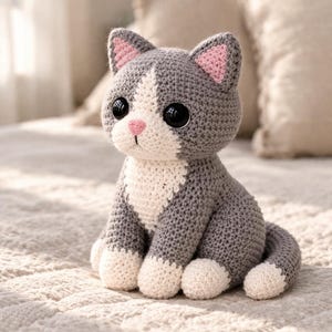 以下が含まれることがあります： グレーと白の手作りの猫の編みぐるみ人形。猫は大きな黒い目、ピンクの内側の耳、ピンクの鼻をしています。人形は質感のある表面に座っており、その詳細な職人技を示しています。