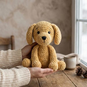 May include: A handmade, golden-yellow crochet puppy dog with floppy ears, a black nose, and black eyes. The plush toy is held in a person's hands, showcasing its soft texture and detailed craftsmanship. The dog is sitting on a wooden surface.