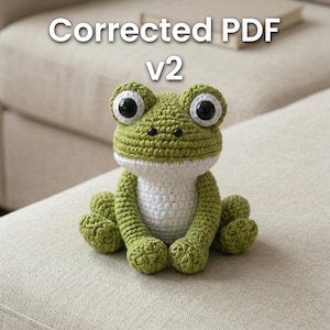 May include: A handmade crochet frog in shades of green and white, with large black and white eyes. The frog is sitting on a neutral-coloured surface. The text "Corrected PDF v2" is displayed above the frog.