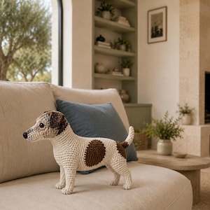 Pode incluir: Uma figura de cão feita à mão em croché com um corpo de cor creme e manchas castanhas na cabeça, costas e cauda. O brinquedo tem um nariz preto, olhos escuros e uma postura de pé com a cauda levantada, exibindo pontos de fio detalhados.