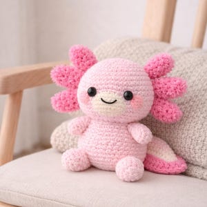 Op de afbeelding: Een handgemaakt roze gehaakt axolotl pluche speelgoed met grote roze franjes, zwarte ogen en een lachend gezicht. Het pluche zit op een beige kussen op een houten stoel. De axolotl heeft een lichtroze staart.