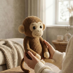 May include: A handmade crochet monkey doll in shades of brown and cream. The monkey has a round head, large ears, and a smiling face. The doll is being held, showcasing its soft texture and detailed craftsmanship. A charming, cuddly toy.