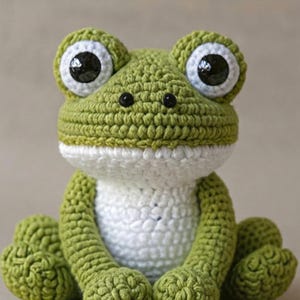 May include: A handcrafted crochet frog in green and white. The frog features large eyes with black pupils and white surrounds. Its body is a mix of green and white, with detailed stitching. A charming, whimsical soft toy.