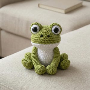 May include: A handmade crochet frog in shades of green and white. The frog has large, expressive eyes with black pupils and white highlights. It is sitting on a light-colored surface, showcasing its detailed craftsmanship. The frog's body is approximately 6 inches tall.