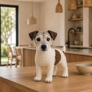 May include: A crochet Jack Russell Terrier dog figurine featuring a white body with brown patches on the side and ears, a speckled gray and white face, black eyes, and a black nose, standing upright with a short tail pointing upward.