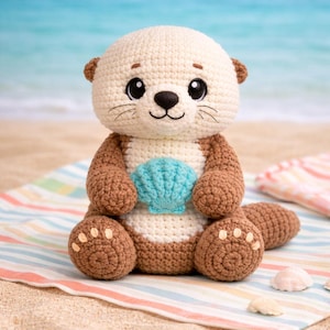 May include: A crocheted otter plush toy with a cream-colored head and a brown body, holding a turquoise seashell. The otter is sitting on a striped blanket on a sandy beach with the ocean in the background.