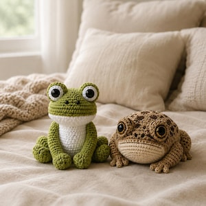 May include: Two crocheted frog plushies. One is green with a white belly, the other is brown with a spotted pattern. Both have large, expressive eyes. They are sitting on a beige bedspread.