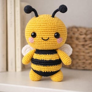 May include: A handmade crochet bee plush toy. The bee is yellow with black stripes, black antennae, and small white wings. It has rosy cheeks and a smiling face. The toy is sitting on a white surface.