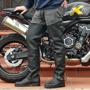 Könnte beinhalten: Ein Paar oberschenkelhohe Stiefel aus schwarzem Leder mit verstellbaren Schnallenriemen am Schaft und am Knöchel. Die Stiefel haben eine glatte, matte Oberfläche, eine abgerundete Zehenpartie und eine dicke Profilsohle in strukturierter Form.