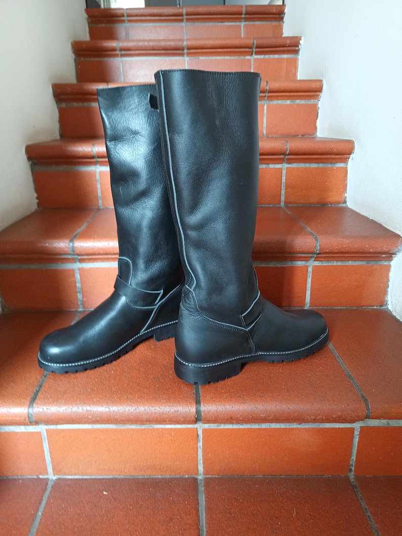 May include: A pair of black leather knee-high boots with a white stitch detail and a buckle on the side.