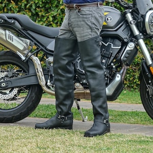 May include: Black leather motorcycle chaps with a buckle detail, worn over gray jeans. The chaps extend from the waist to the ankles. A motorcycle is in the background.