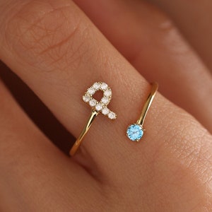 Può includere: Un anello aperto in oro con un piccolo ciondolo a forma di nastro incastonato con minuscole pietre chiare su un'estremità e una singola pietra preziosa rotonda azzurra sull'altra. La fascia è sottile e lucida, per un aspetto delicato e raffinato.