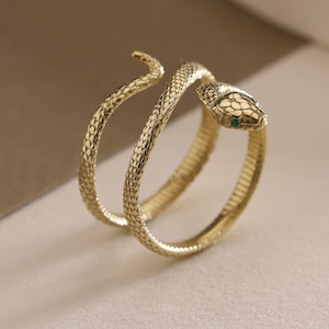 May include: A gold-colored snake ring with a textured, scale-like pattern. The snake's head features a small green stone. The ring wraps around the finger in a spiral design.