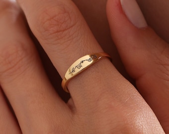 May include: A gold band ring with a delicate floral design engraved on the top. The ring is worn on the middle finger of a hand.