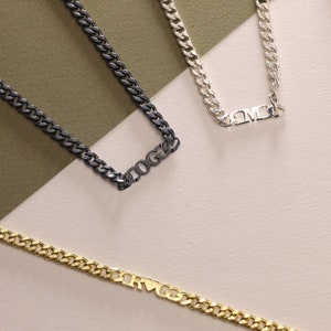 Könnte beinhalten: Drei goldene Kettenhalsbänder mit verschiedenen Anhängern. Die erste Halskette hat einen schwarzen Anhänger mit dem Wort "TOGA" darauf. Die zweite Halskette hat einen silbernen Anhänger mit den Buchstaben "DMC" darauf. Die dritte Halskette hat einen goldenen Anhänger mit den Buchstaben "RVGG" darauf.